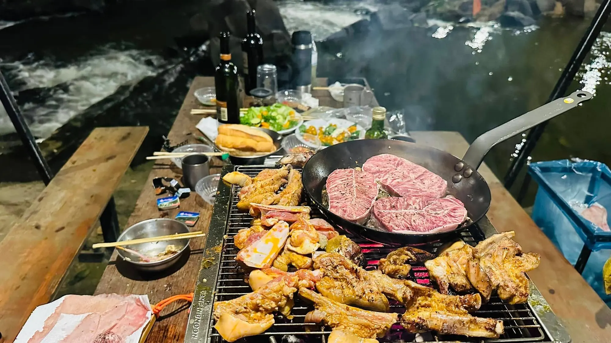 शाम को धारा किनारे मांस, सॉसेज और बीफ़ के पैन के साथ BBQ ग्रिल