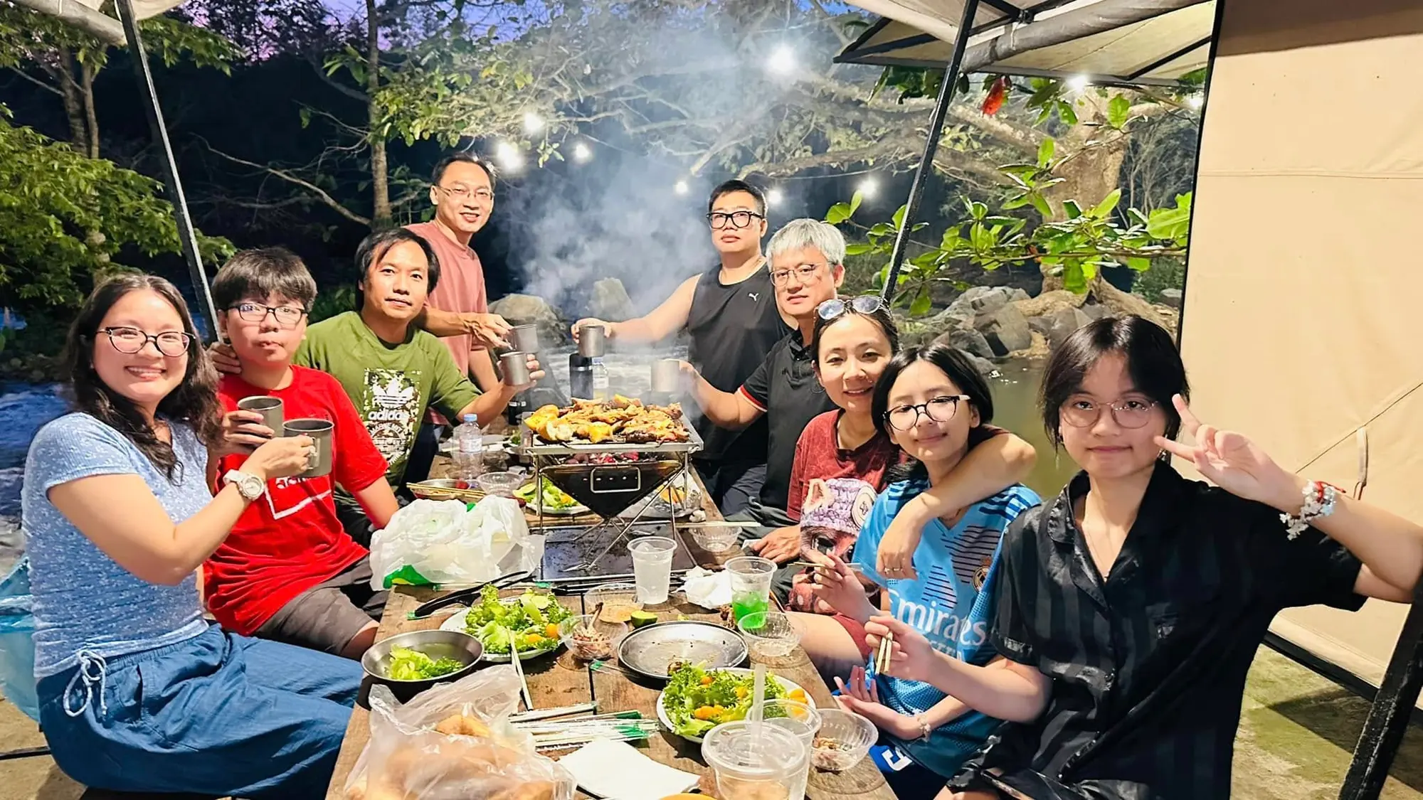 शाम को BBQ टेबल के चारों ओर इकट्ठा मेहमानों का समूह, ग्रिल किए व्यंजनों के साथ हँसते-मुस्कुराते
