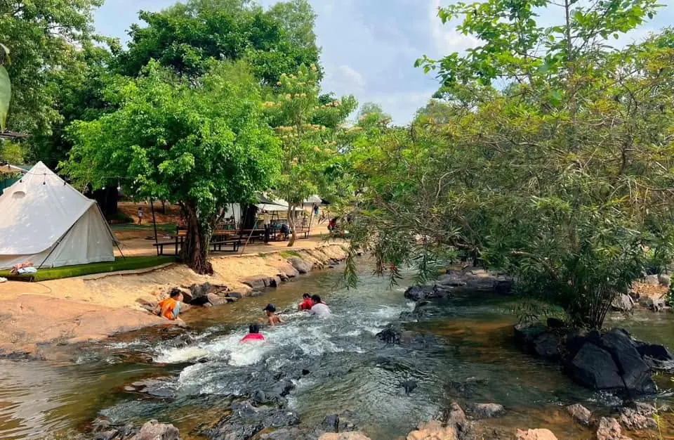 चट्टानों के बीच धारा में नहाते मेहमान, किनारे पर पेड़ों की छाँव में सफ़ेद घंटीनुमा टेंट